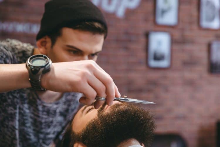 Young consentrated barber grooming beard of man with scissors